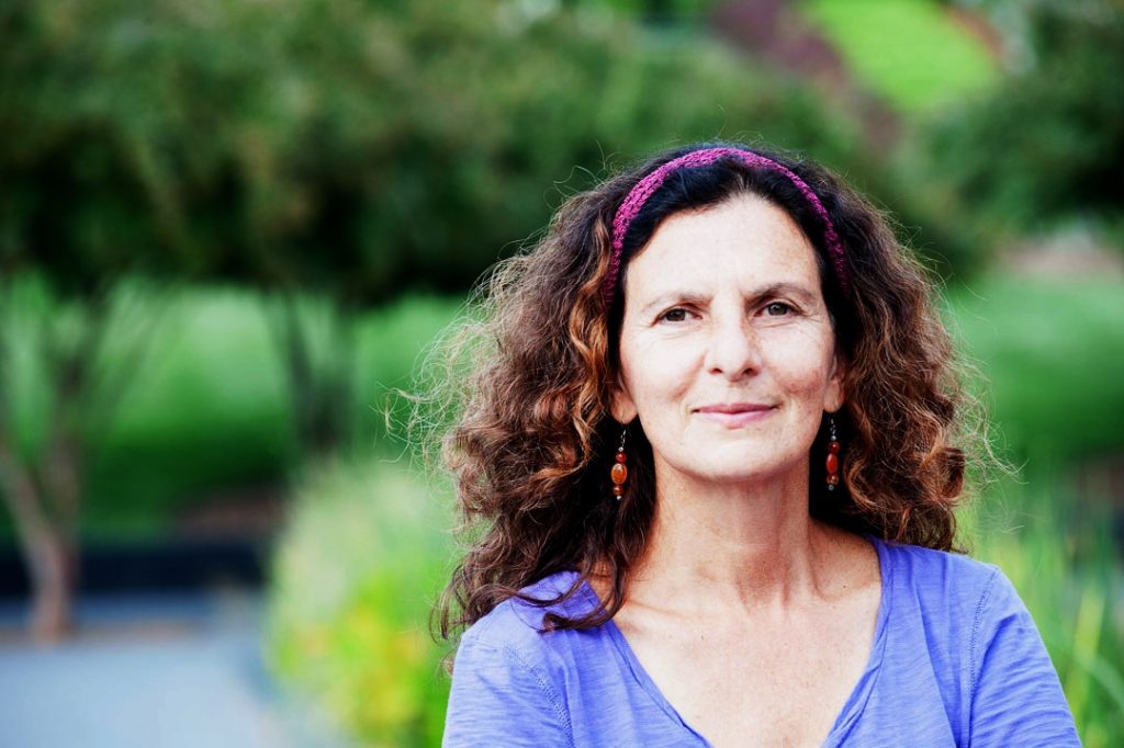 Image: Photo of Author, Poet, Dancer, and Teacher Cheryl Pallant. Ms. Pallant is facing the camera, smiling. She is wearing a purple shirt, her hair long, wavy, and flowing. In the background, a beautiful, green, natural setting. 