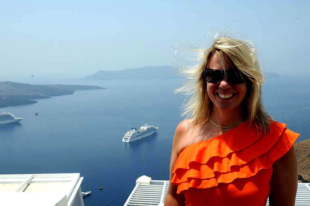 Award-winning thriller author K.J. Howe, creator of the Thea Paris series. 

A photo of Ms. Howe in Santorini, wearing an orange shirt. She is standing to the right of the frame, with the Aegean Sea behind her and a yacht off in the distance.