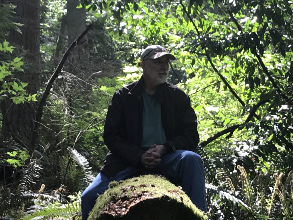 Dr. Mark Seely, author of Stones: Meditations on Human Authenticity, sitting on a log in Southwest County Park